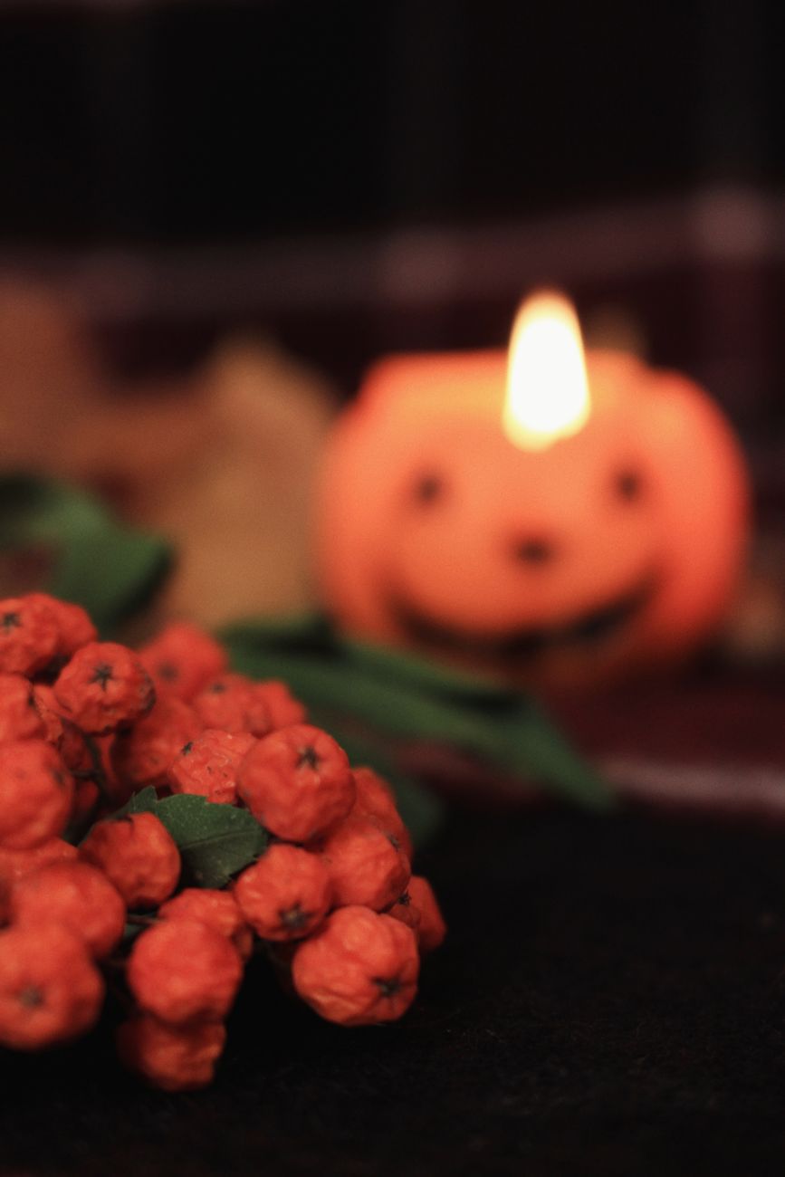 A close-up of a cluster of small orange berries with green leaves, with a blurred, smiling jack-o'-lantern candle in the background.